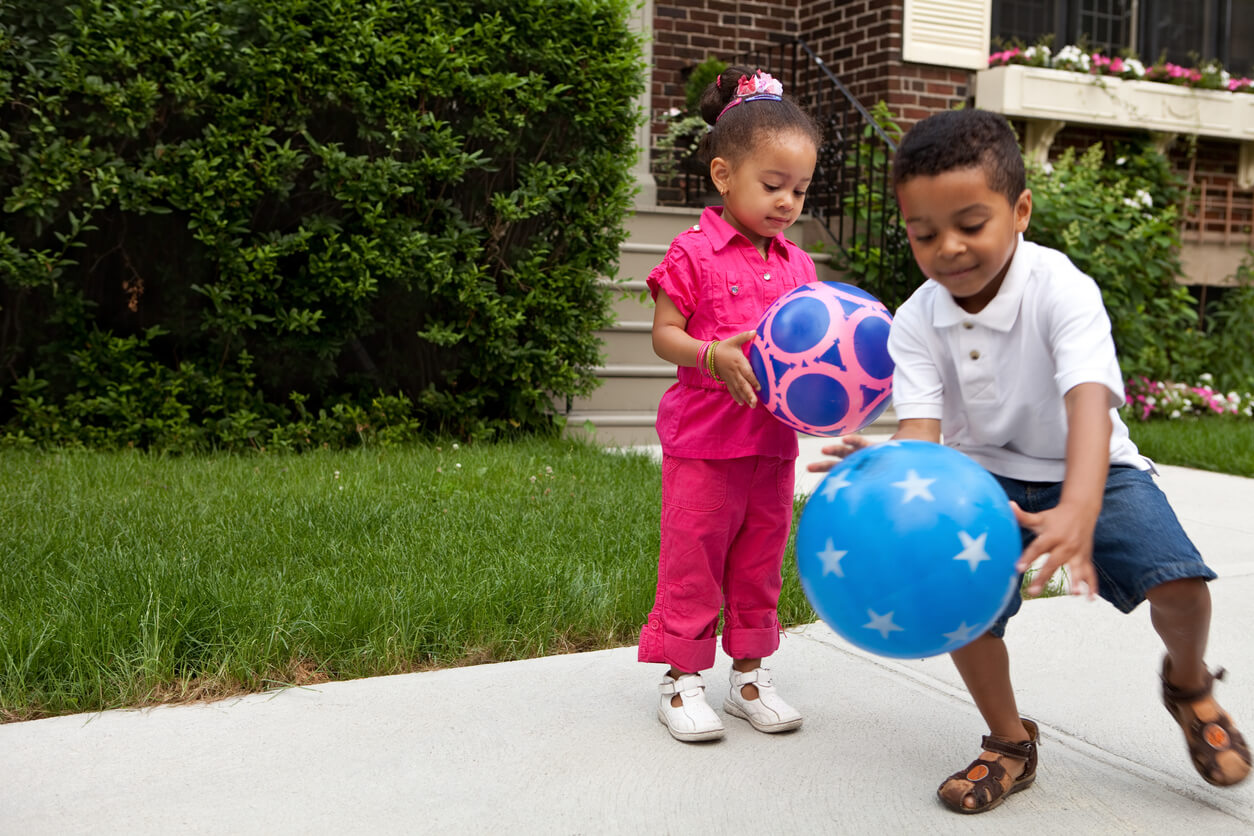 A brother and sister playing with a bouncy ball on the sidewalk.