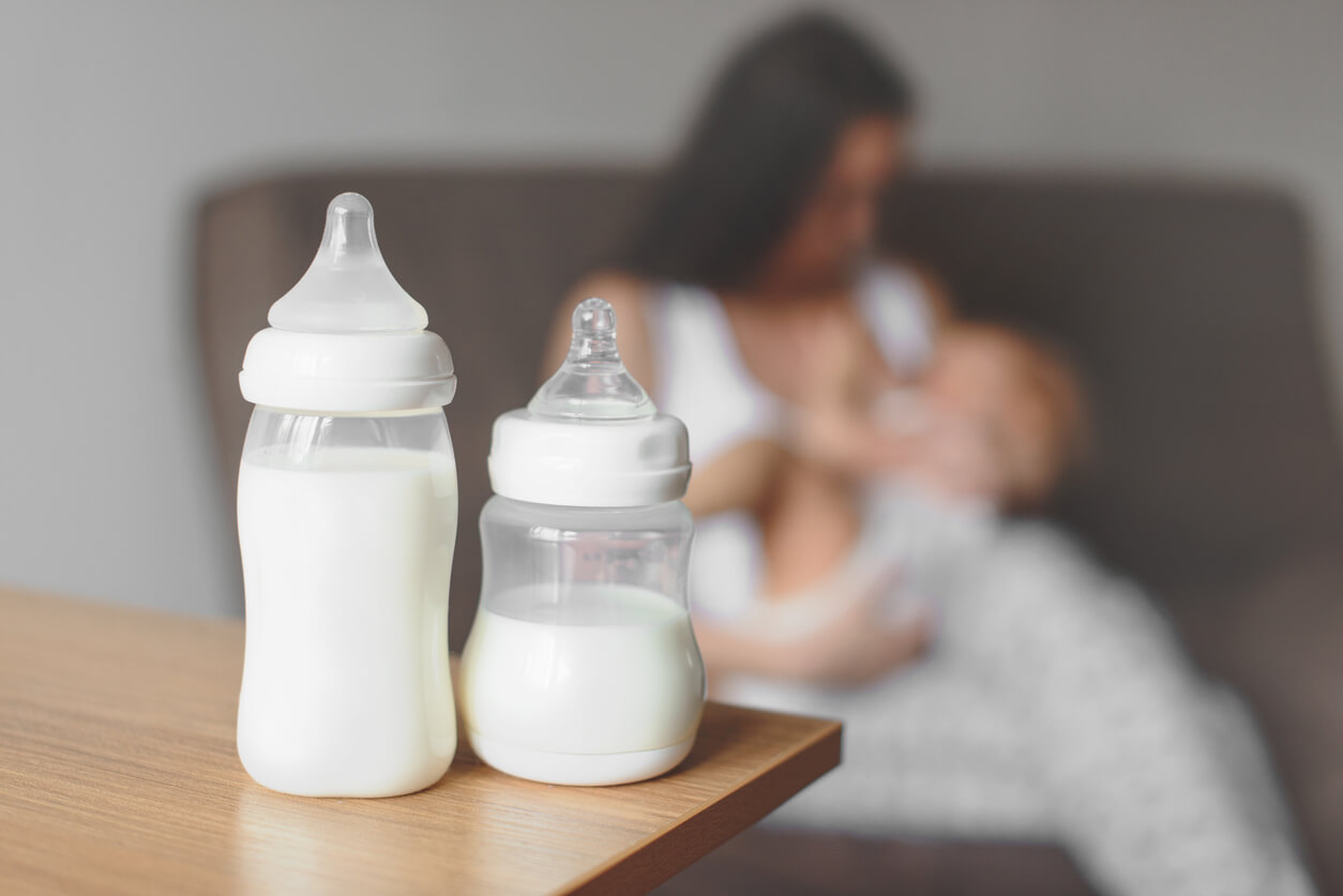 Two baby bottles of milk sitting on a table while a mother breastfeeds her baby in the background.