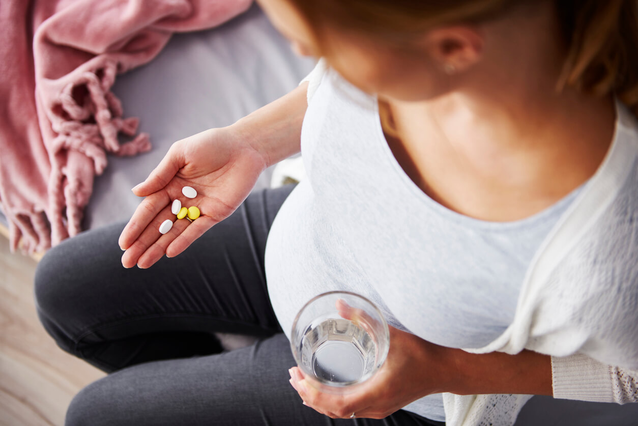 A pregnant woman holding a glass of water in one hand and 5 pills in the other.