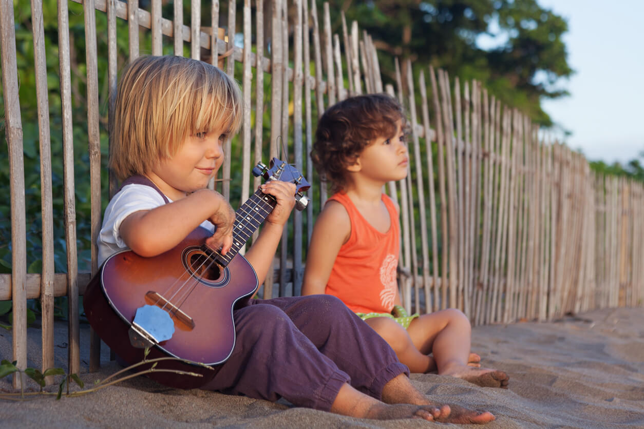 To små gutter som sitter på stranden mens en av dem spiller ukelele.