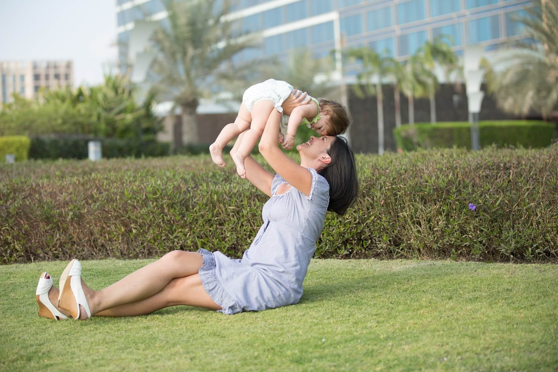 En mor sitter i gräset och lyfter sin bebis i luften.