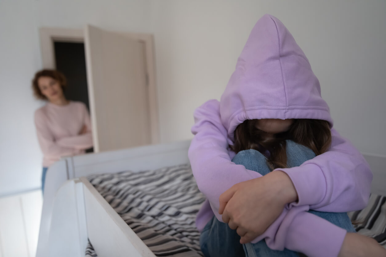 A teenager sitting in bed looking upset, with her back to her mother.