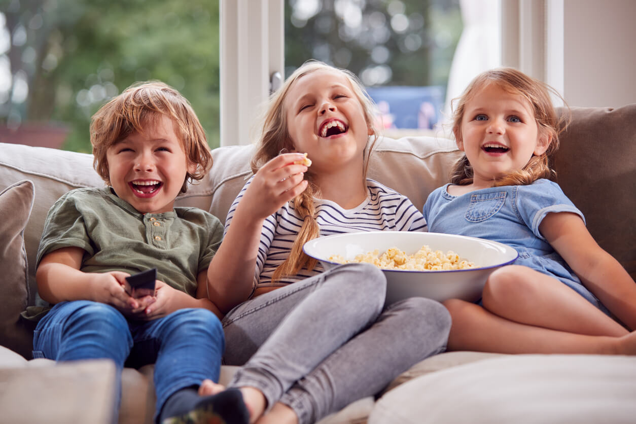 Young siblings eating popcorn and laughing while watching TV on the couch.