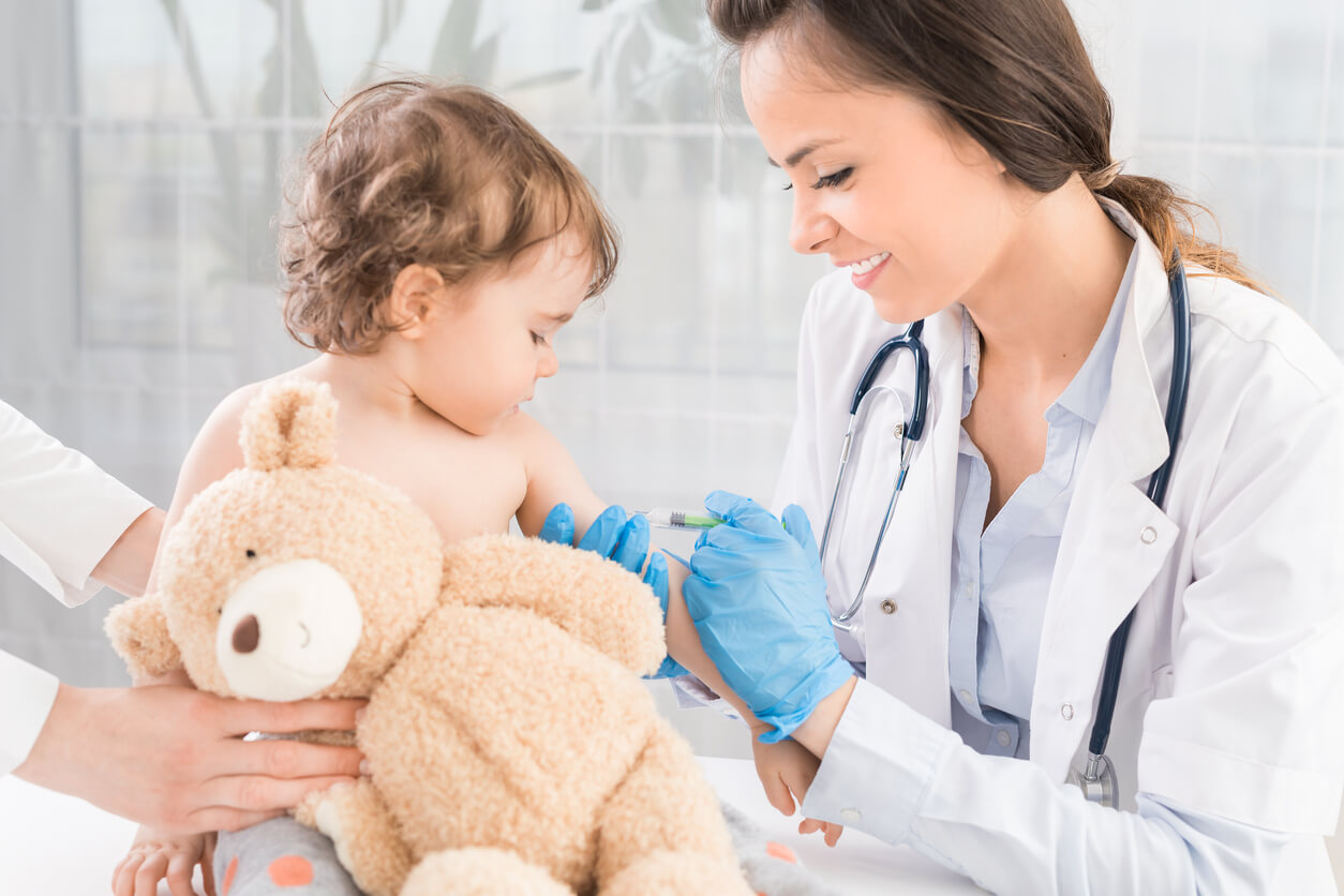 A nurse giving a baby a vaccine while she holds a teddy bear.