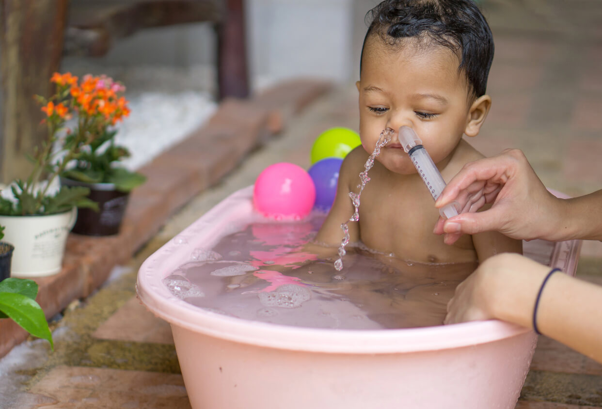A woman performing a nasal wash on a baby who's sitting in a bathtub.
