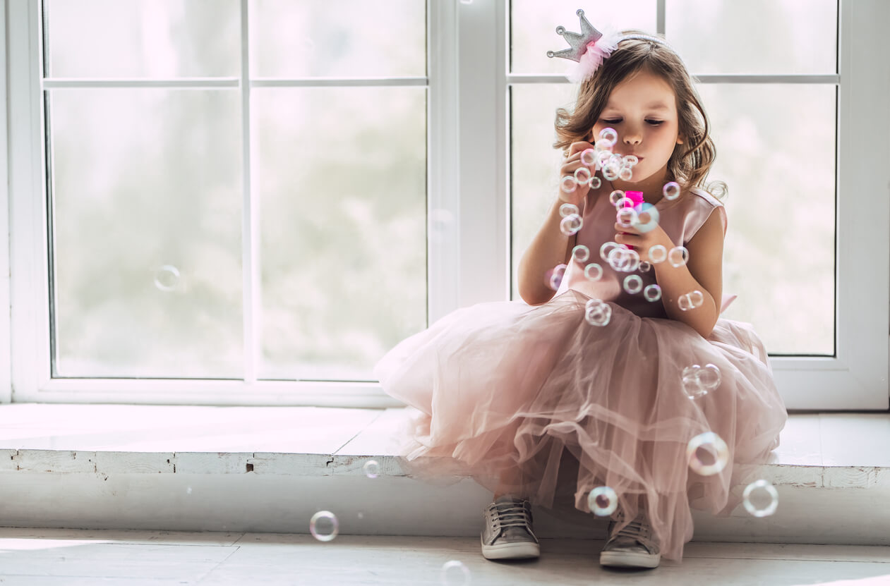 A young girl dressed as a princess, sitting by a window, blowing bubbles.