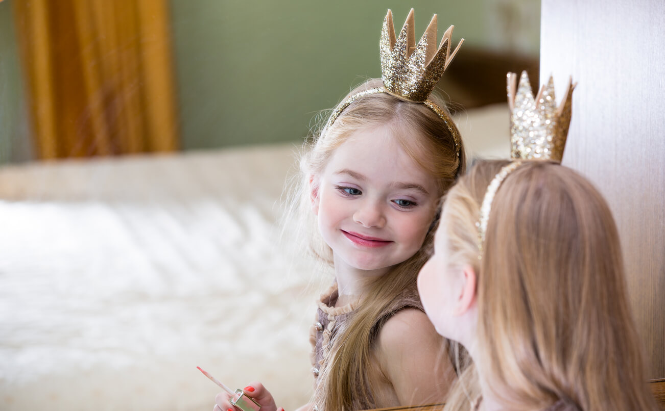 A little girl wearing a crown and smiling at herself in the mirror.