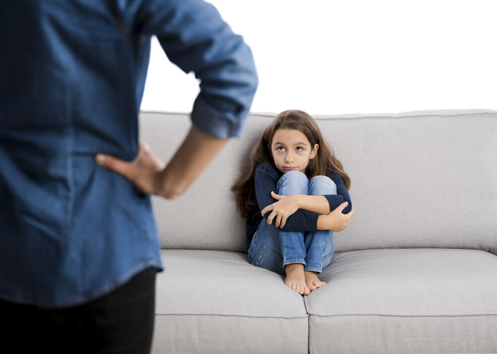 A little girl sitting on a couch looking guilty as her mother looks at her with her hands on her hips.