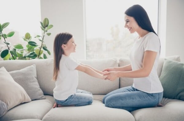 A mother and daughter sitting on the couch facing one another and holding hands.
