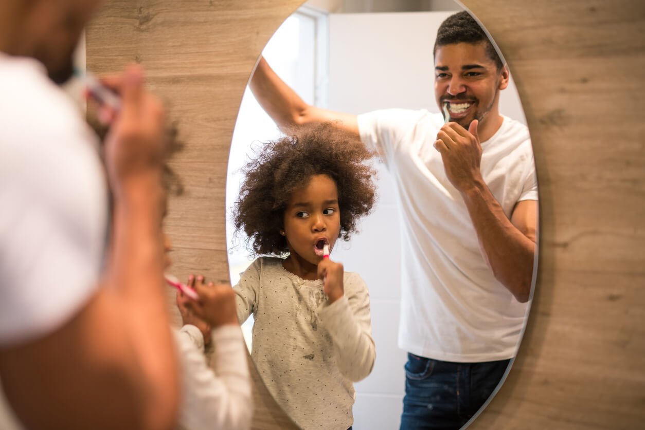A father and daughter brushing their teeth together in front of the mirror.