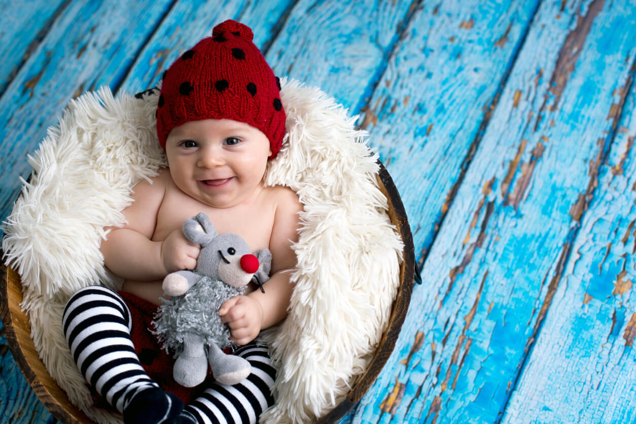 A baby boy wearing a red knit cap, black and white stckings, and holding a stuffed mouse.
