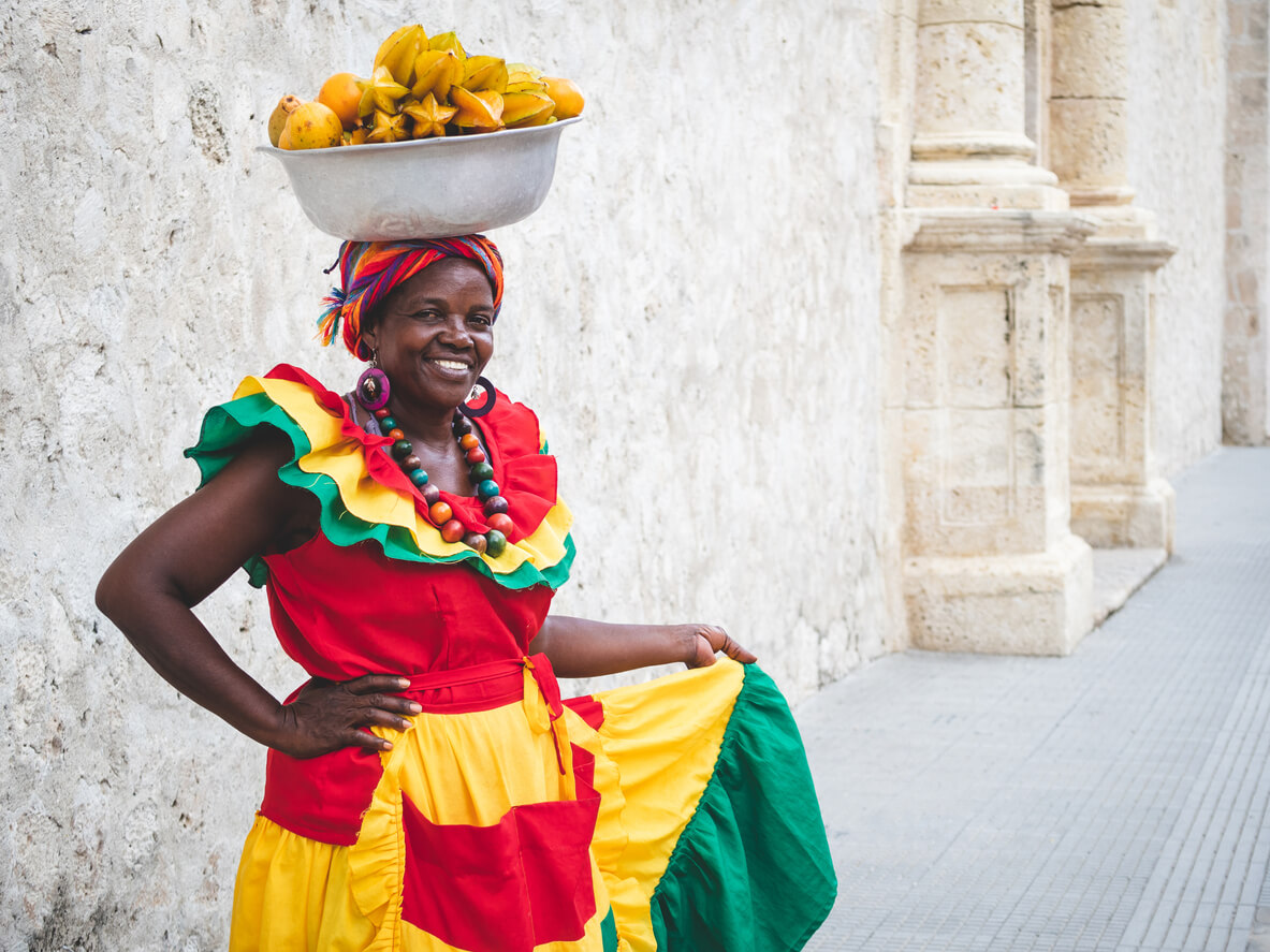 A Columbian woman wearing traditional dress and carrying a bowl of fruit on her head.