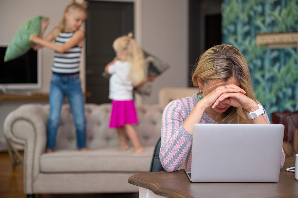 A mother having a hard time concentrating on her work on the computer while her daughters have a pillow fight on the couch.