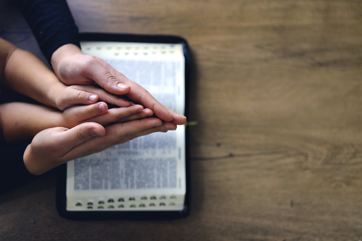 Mãos de mulher e criança na Bíblia em posição de oração.