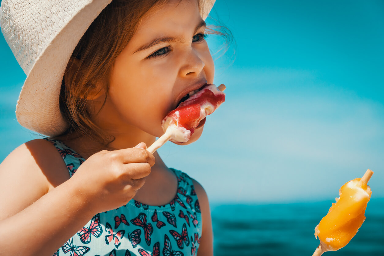 menina come sorvete de frutas na praia mar chapéu maiô sol