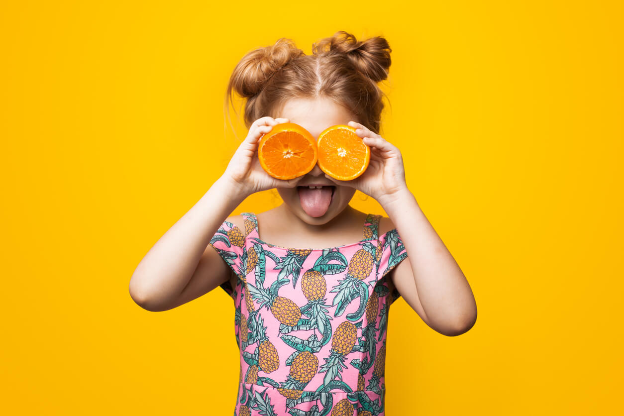 A little firl wearing a pineapple print shirt holding two orange halves in front of her eyes and sticking out her tongue.