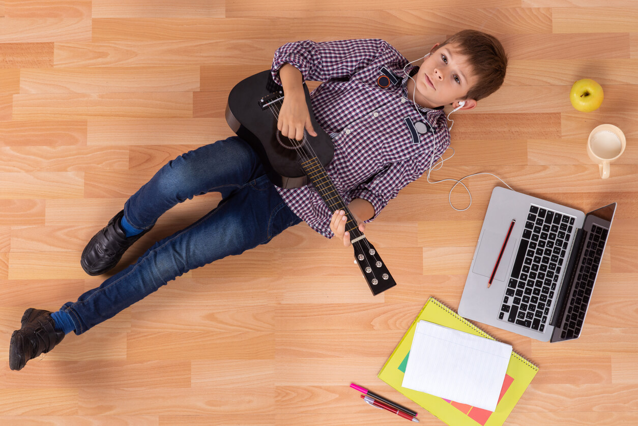 menino em um momento de distração no estudo com seu violão no chão fones de ouvido laptop folhas caderno