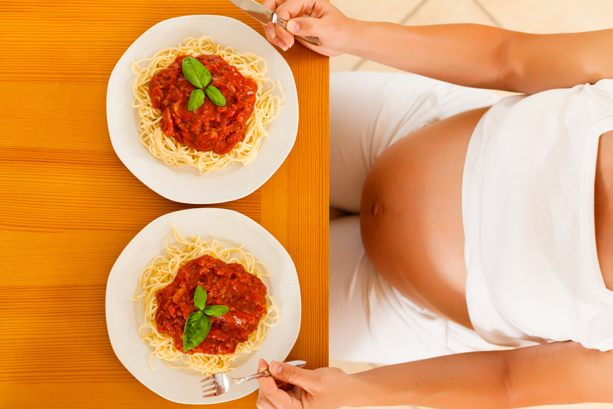 A pregnant woman sitting down in front of two plates of pasta.