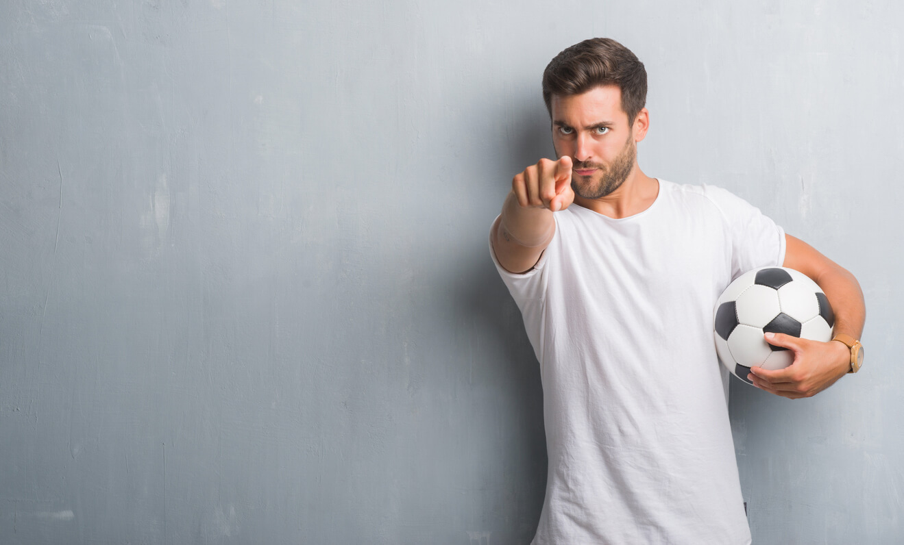A soccer coach holding a ball and pointing at the camera.