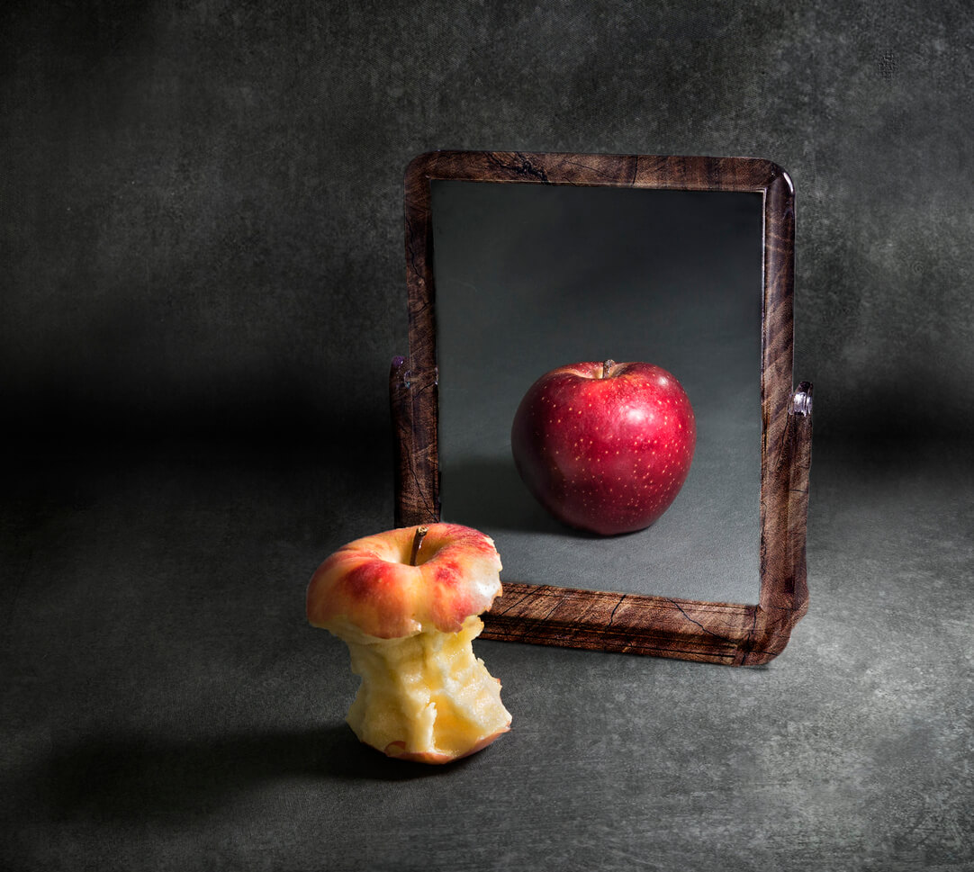 An apple core that's reflected in a mirror as an uneaten apple.