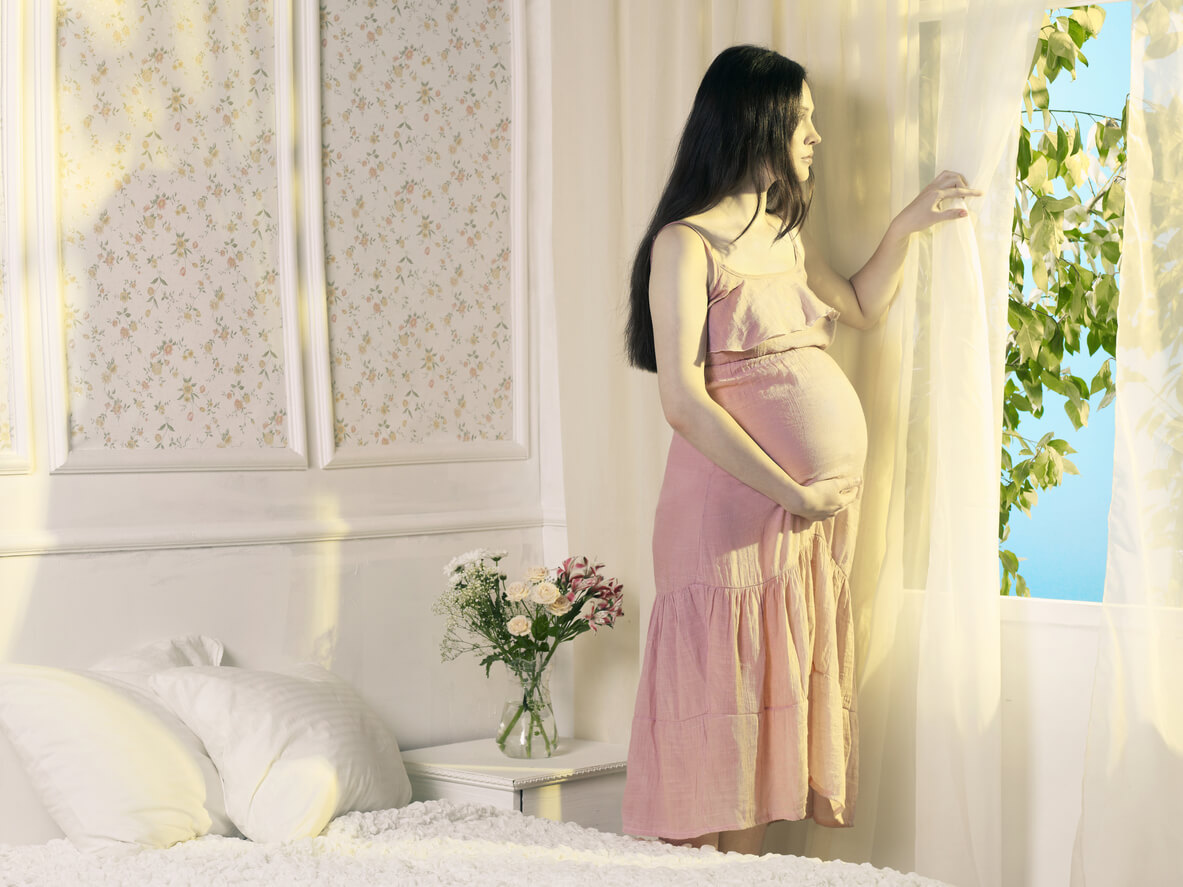A pregnant woman looking out the window during summer.