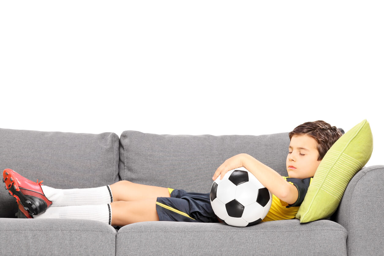 A child sleeping on the couch in his soccer jersey while holding a soccer ball.