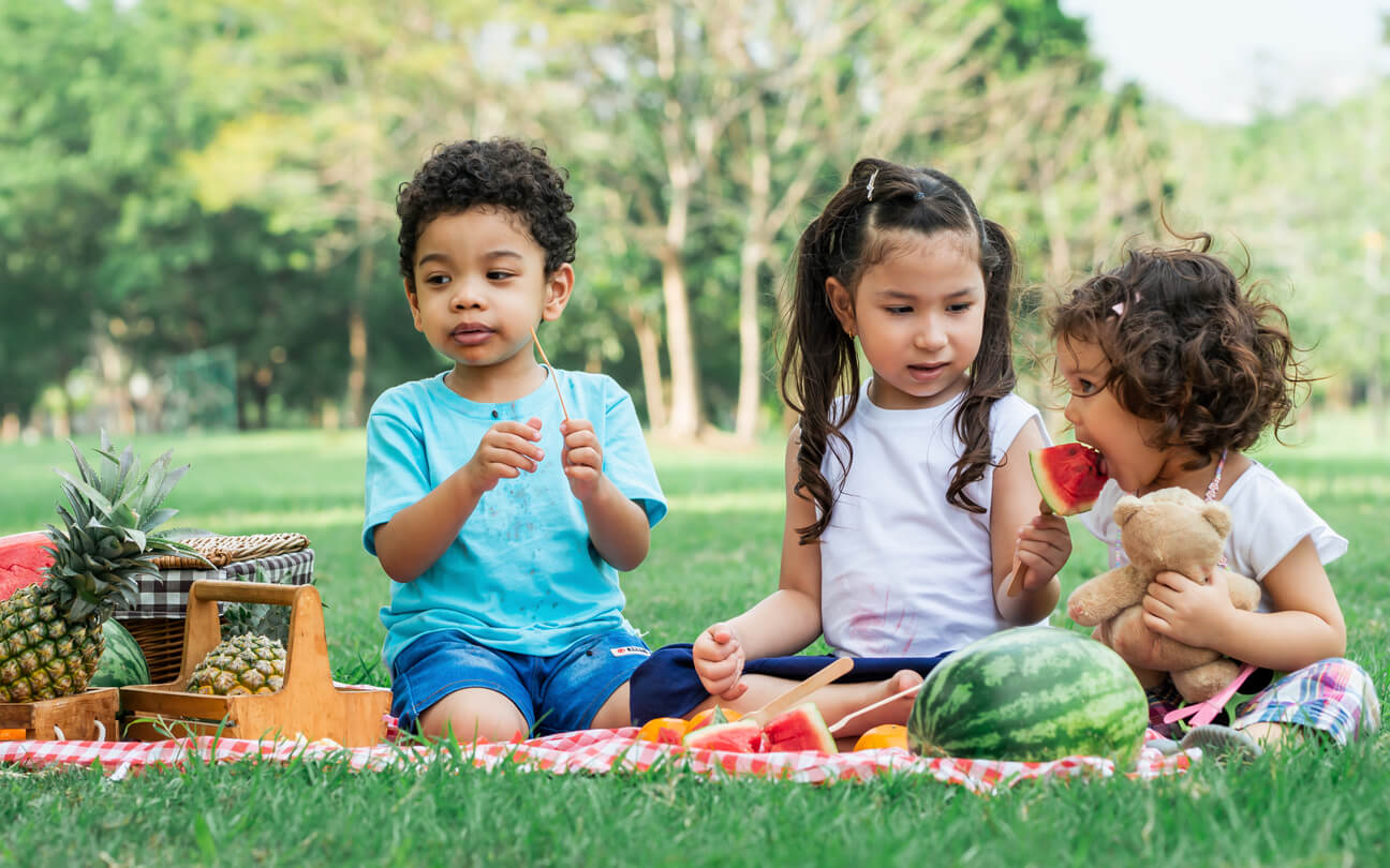 Children eating watermelon on a picnic blanket in the grass.