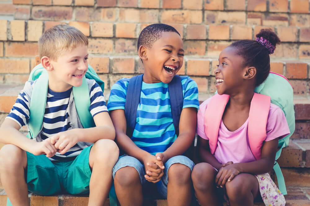To svarte barn og et hvitt barn sitter og ler sammen.