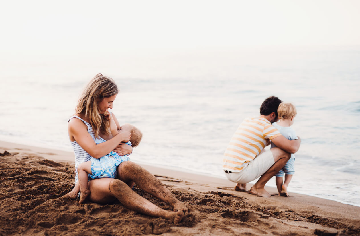 A mother breastfeeding her child on the beach while her parther plays with their other child.