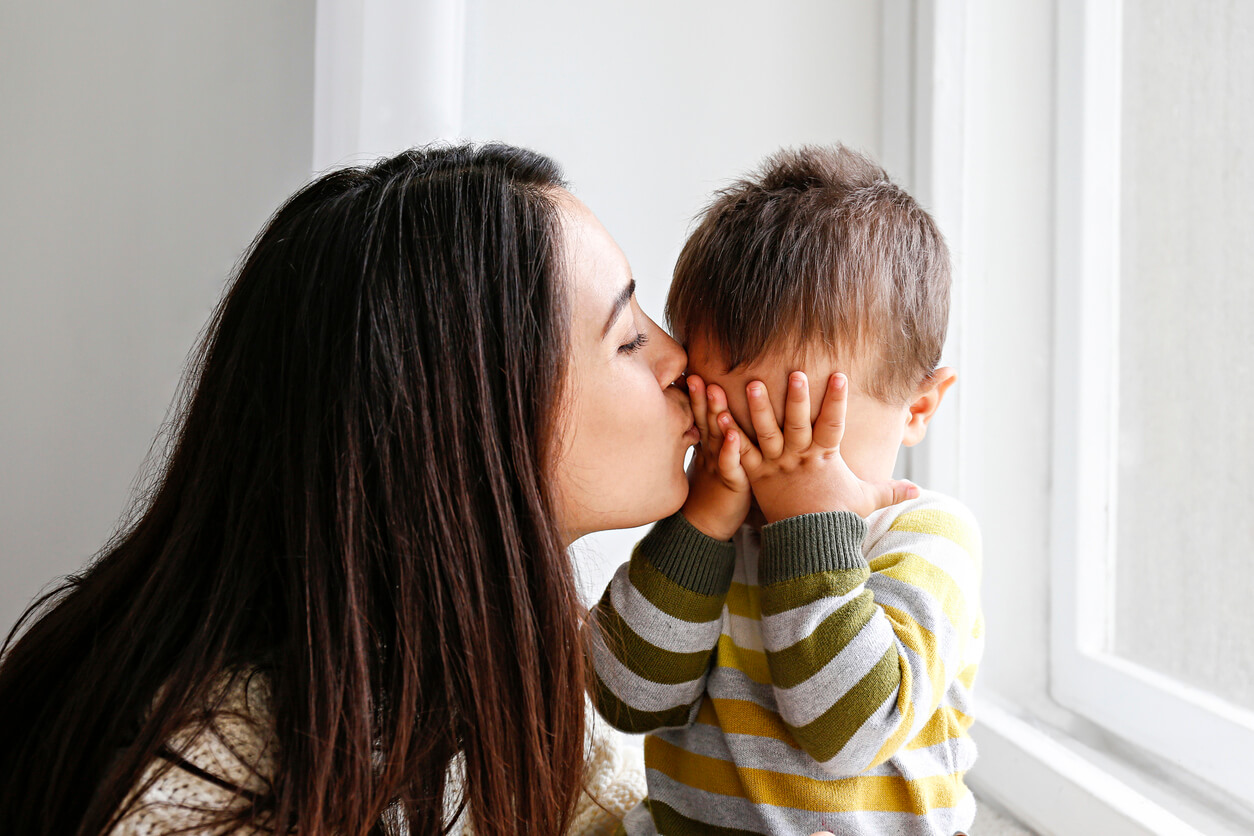 En bebis som täcker ögonen med händerna samtidigt som han får en kyss från sin mamma.