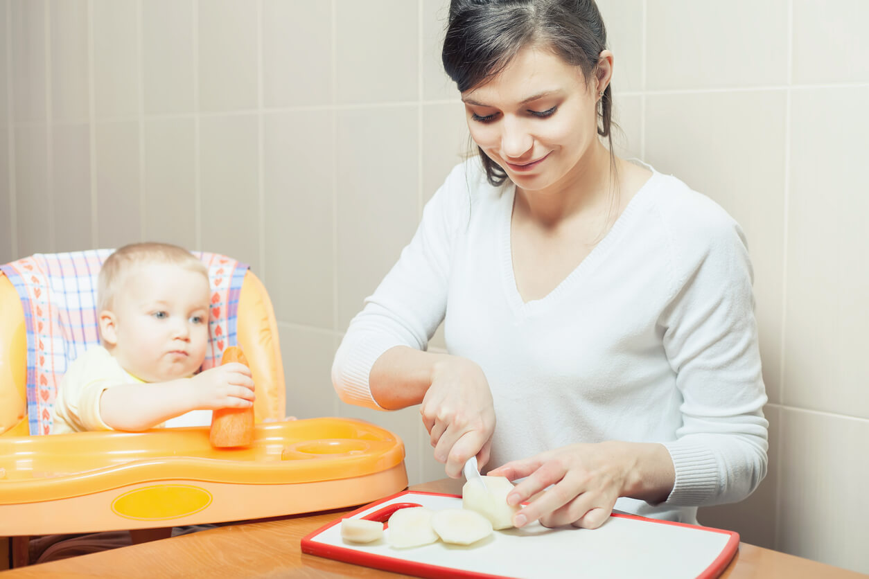 A baby holding a carrot while his mother cuts an apple.