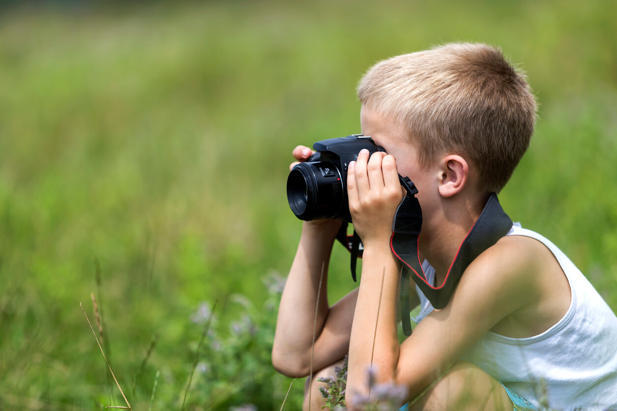 A young boy sitting in a field looking through a camara.
