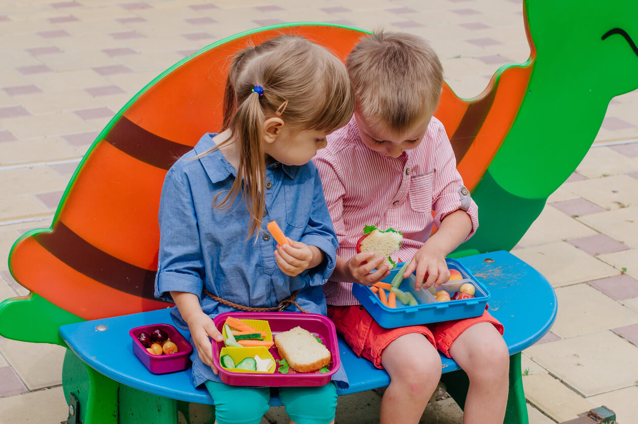 Two preschoolers eating lunch together on a bench.