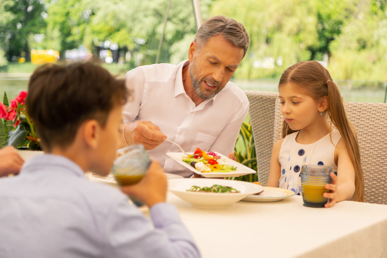 A father serving vegetables to his daughter as they dine outdoors with their family.