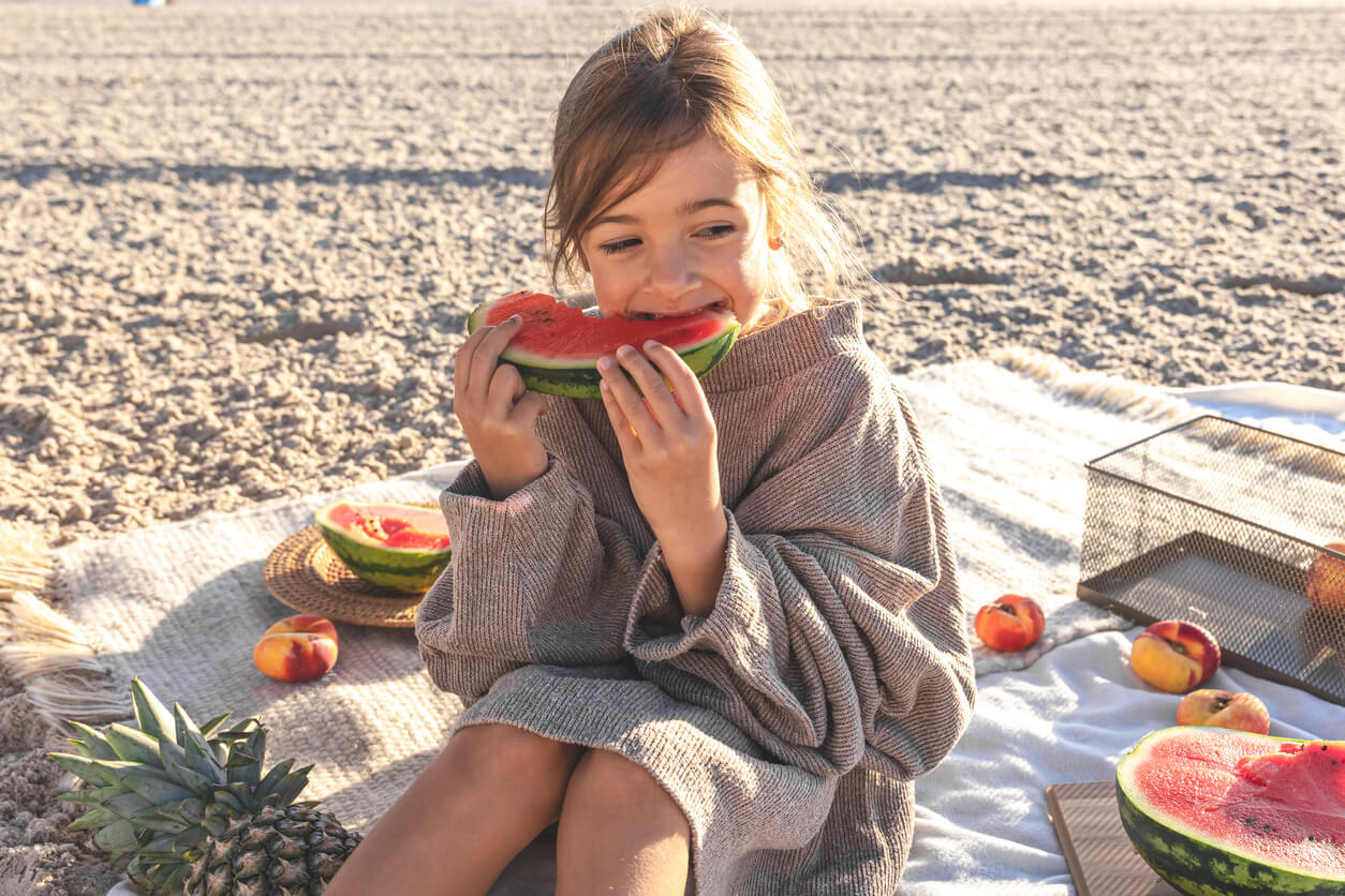 A little girl sitting on a blanket in the sand, eating watermelon.
