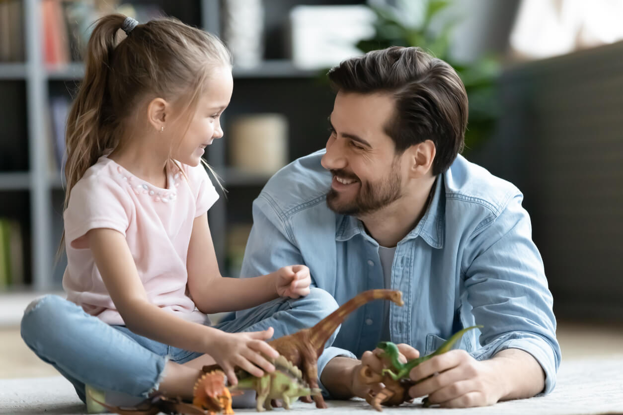 A little girl sitting on the floor with her dad, playing with dinosaurs.