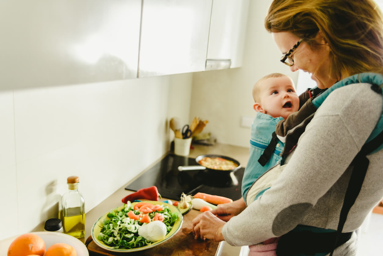 A woman smiling at her baby in a baby carrier while she prepares a salad for herself.