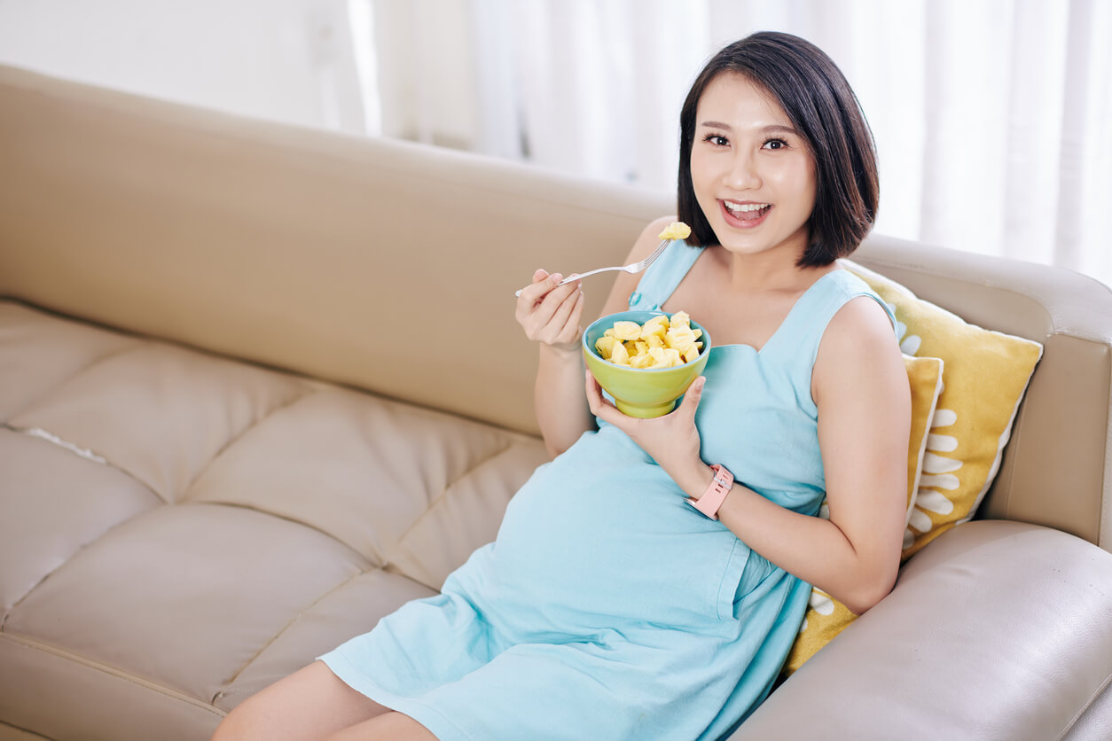 A pregnant woman sitting on the couch and eating pineapple.