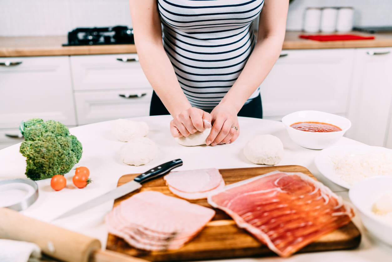 En gravid kvinne som forbereder seg på å lage med deig, skinke, grønnsaker og andre ingredienser.