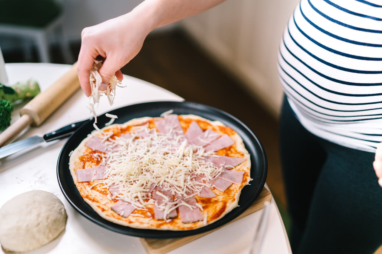 A pregnant woman preparing a ham and cheese pizza.