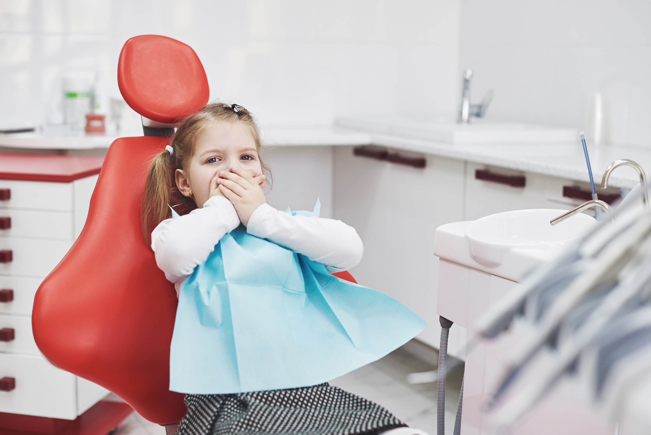 A little girl sitting in a dentist's chair covering her mouth with both hands.