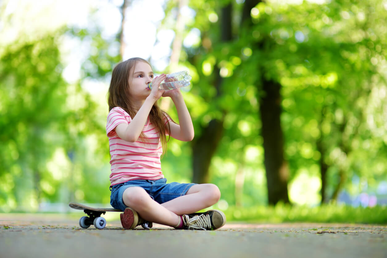 A young girl sitting on a skateboard and drinking water in the park.