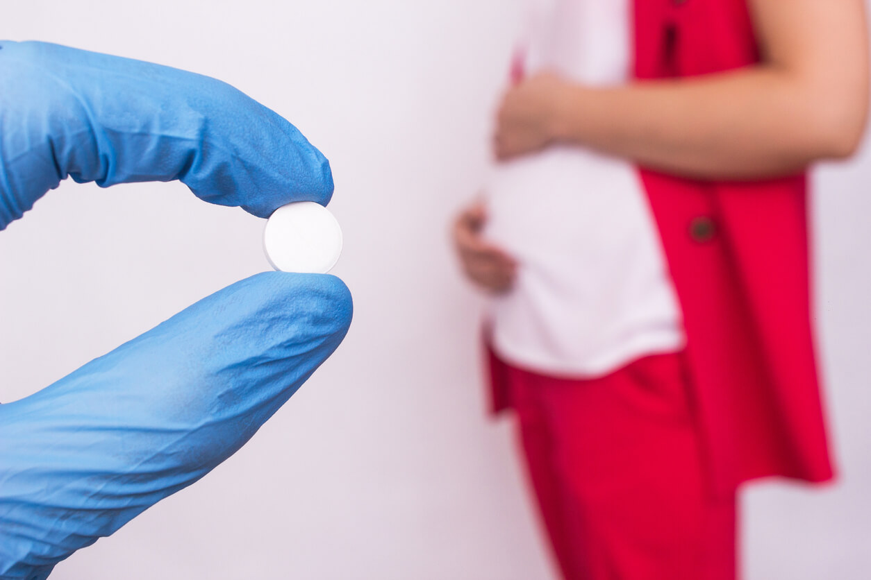 En handskförsedd hand som håller ett piller och den suddiga bilden av en gravid kvinna i bakgrunden.
