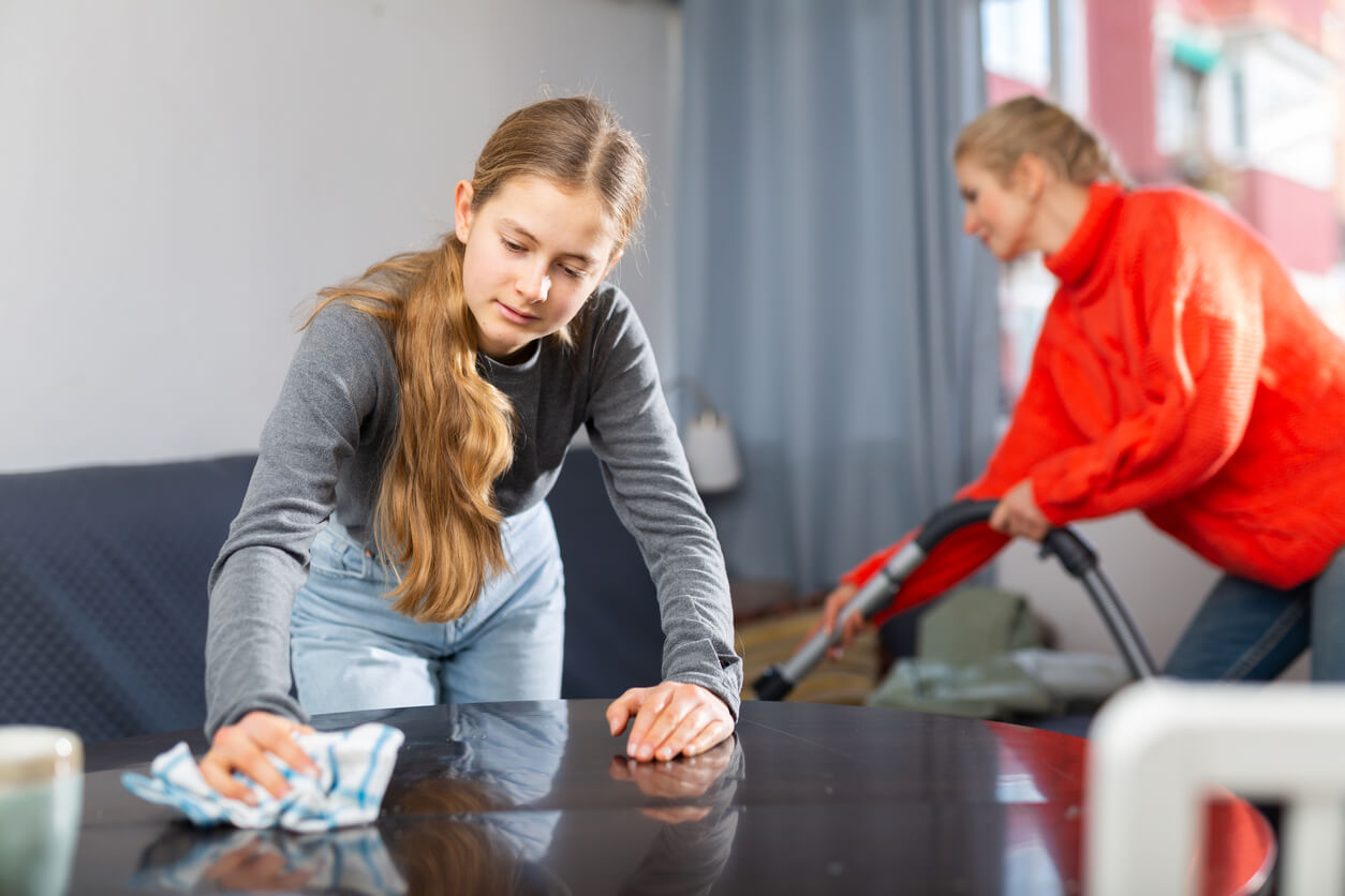 A teen girl cleaning the table while her mother vacuums.
