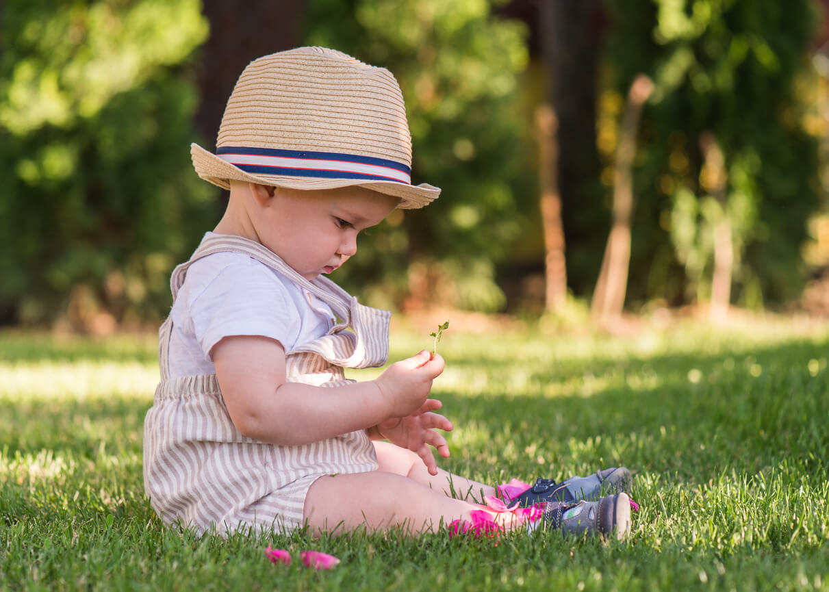 Vauva istuu nurmikolla hattu yllään ja leikkii kukilla.