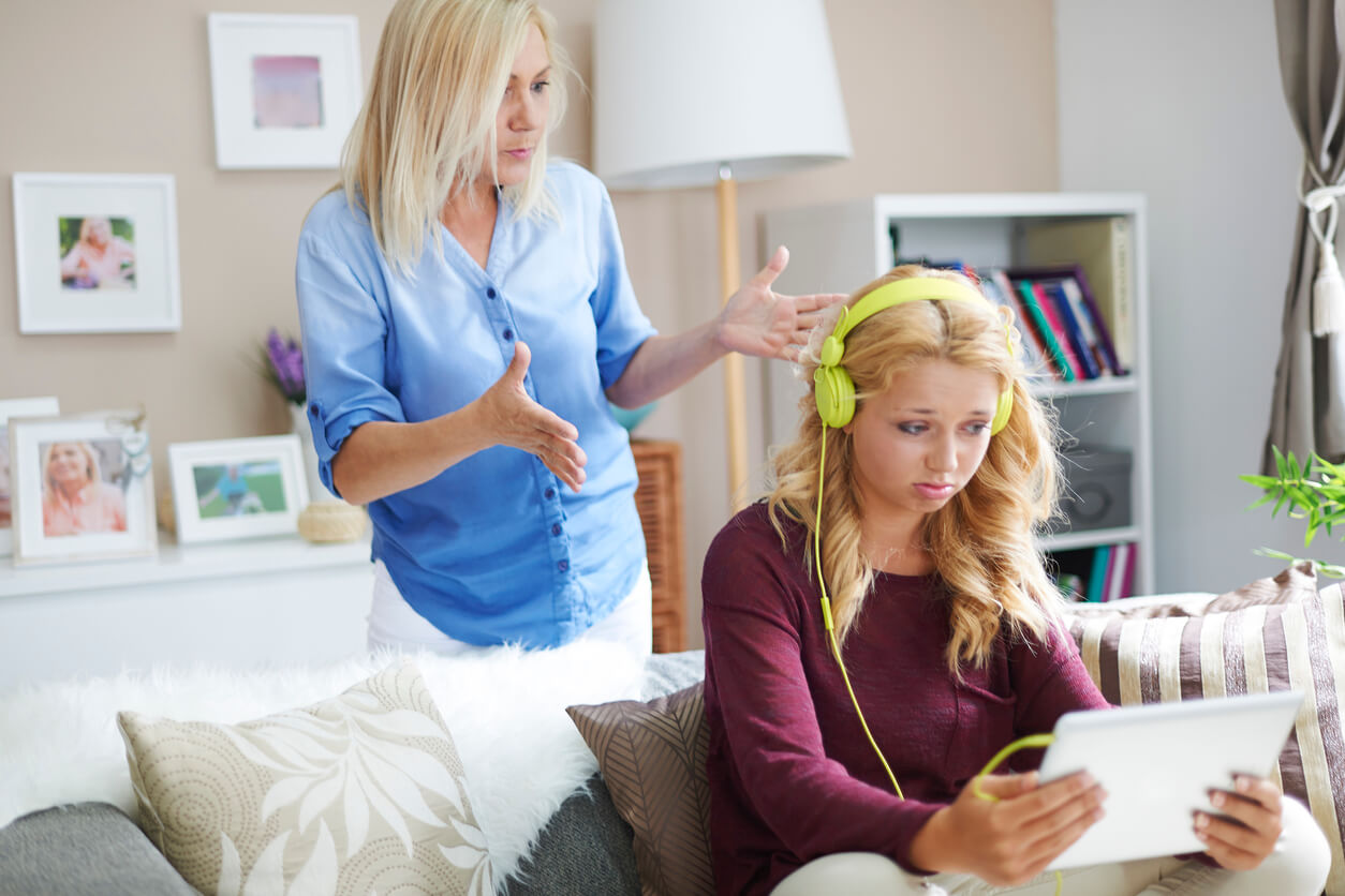 A mother who looks annoyed while her teen daughter looks at her tablet and listens to headphones.
