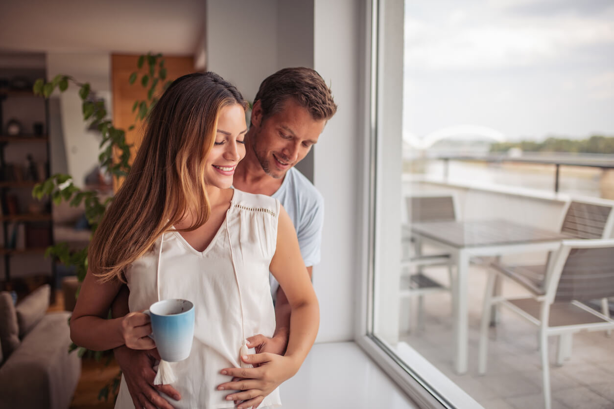 A pregnant woman smiling as her partner stands behind her with his hands on her belly.