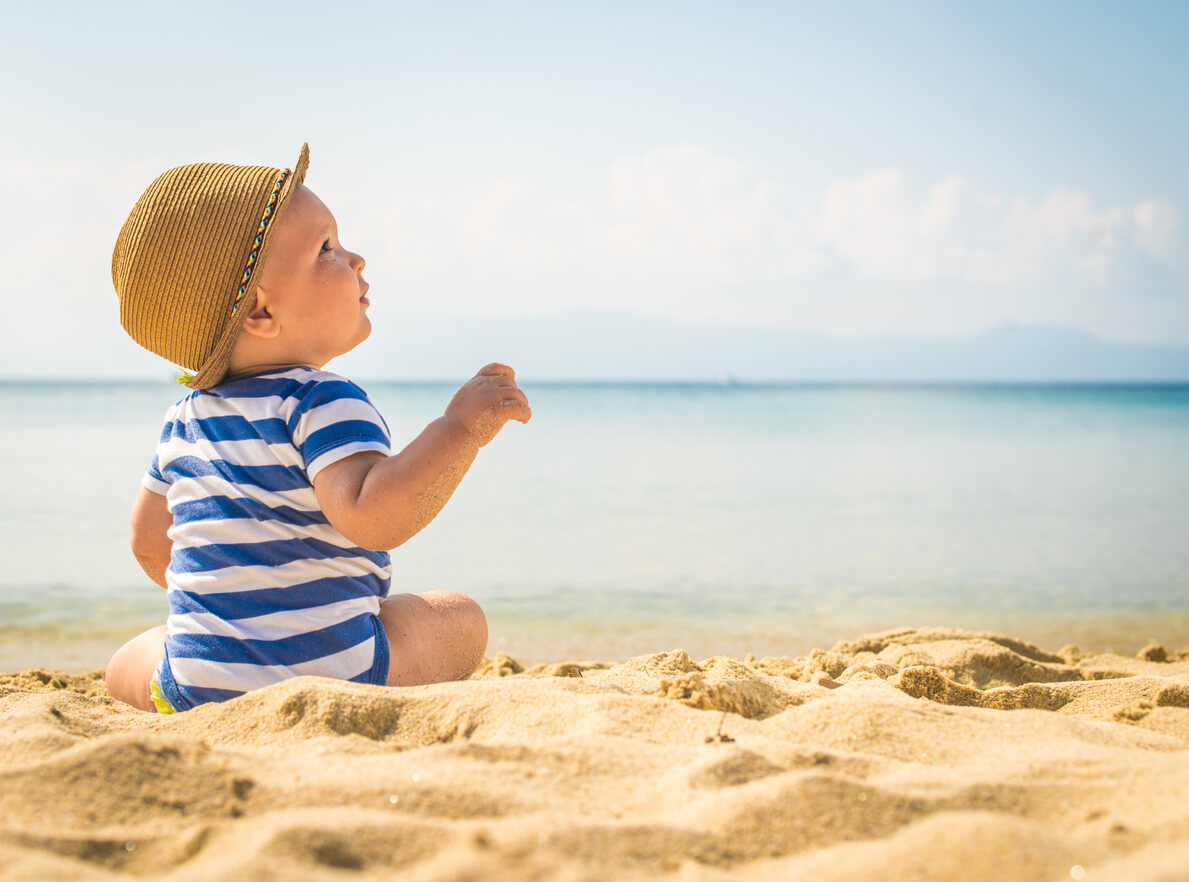 A baby on sitting on the sand without sunglasses.