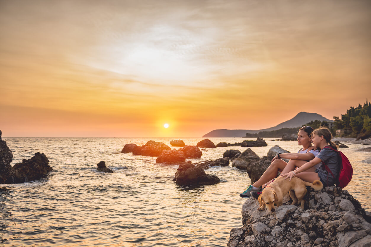 En mor, datter og hunden deres sitter på en stein og ser ut over havet.