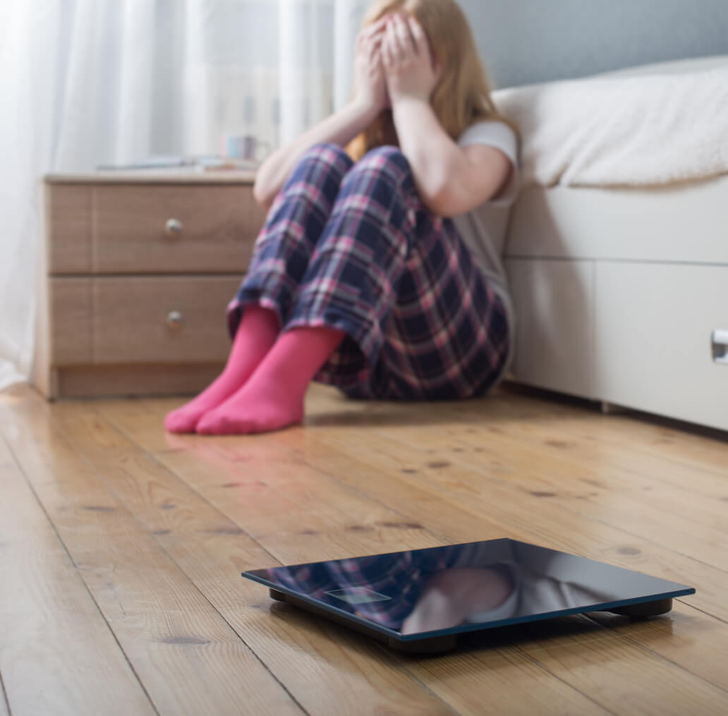 En tenåringsjente som sitter på gulvet og dekker ansiktet hennes, med en tom tallerken i forgrunnen.